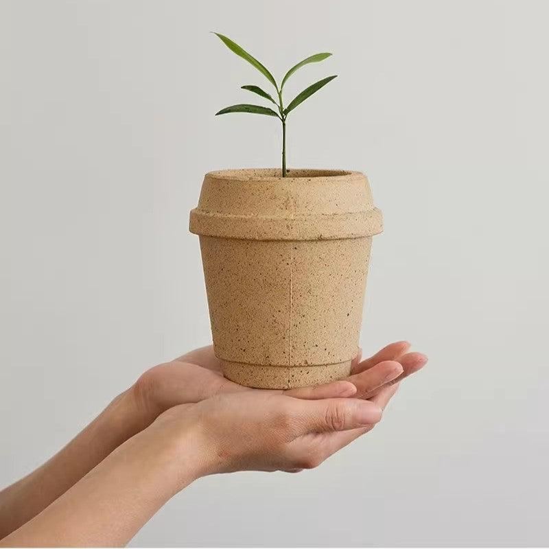 4 inch Natural-colored ceramic coffee cup-shaped planter with a young seedling, held in open hands, showcasing a minimalist, eco-friendly design.