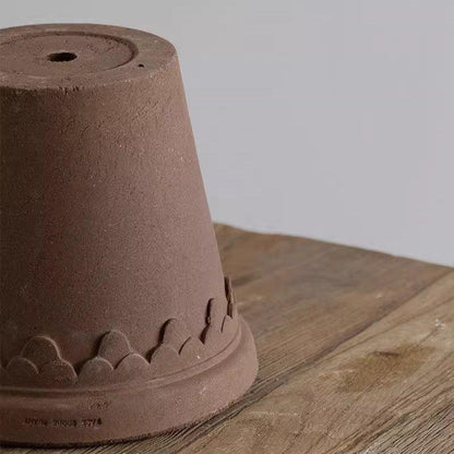 Chocolate brown clay planter with a drain hole and decorative scalloped design, positioned upside down on a rustic wooden surface.