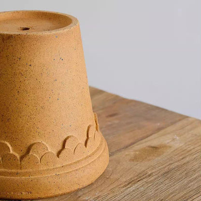 Ochre clay plant pot with drainage hole and scalloped accents, showcasing a natural speckled texture, placed upside down on a wooden surface.